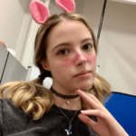 A white female with blonde hair in two side ponytails wearing pink upright bunny ears, pink makeup and a black choker, posing for the camera