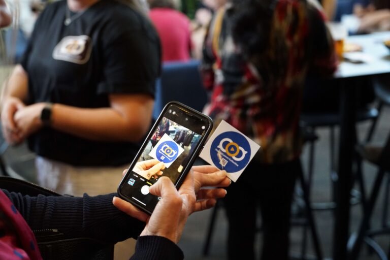 Someone scanning a QR code on a mobile phone. The QR code is part of a round blue sticker that is advertising the Sci Comm Collective.