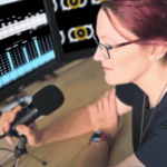 White woman with dyed red hair at a desk microphone, a screen with audio editing and another with the podcast logo on it before her