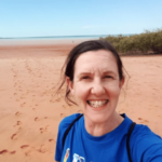 a smiling white woman with a red sand beach and blue sky behind her. She is wearing a blue shirt.