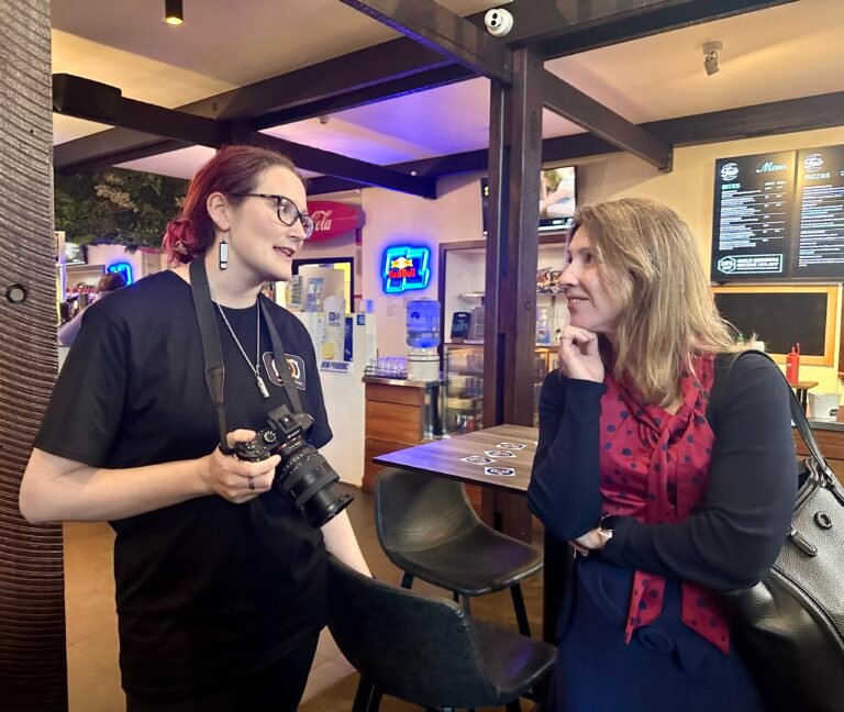 Two white women engaging in conversation with each other. One has dark red hair and has a camera around her neck. The other is resting her chin on her hand and looking at the other woman.