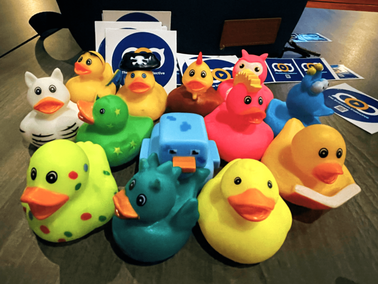 A dozen brightly coloured rubber ducks on a table. They are surrounded by blue stickers with The Sci Comm Collective logo on them.