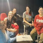 a group of five people of various ages and background sitting around a table in a recording studio. They are smiling at the camera.