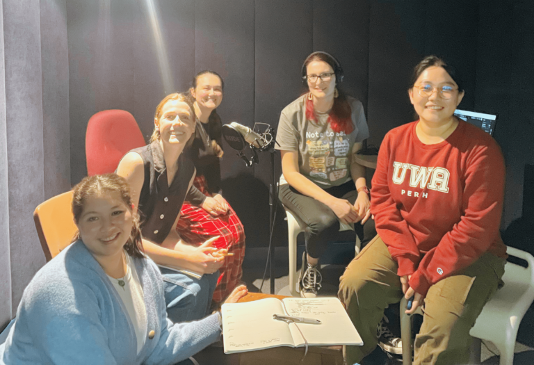 a group of five people of various ages and background sitting around a table in a recording studio. They are smiling at the camera.