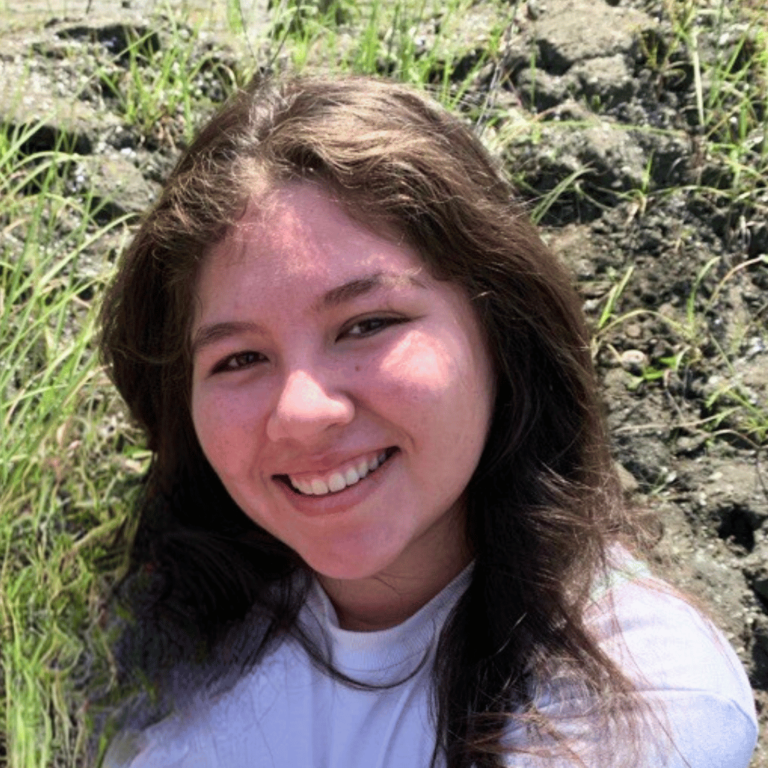 young Central American woman with brown hair smiling at the camera, grass and rocks behind her