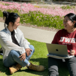 two people sitting on the grass with pink Western Australian wildflowers in the background. One person is wearing a maroon UWA jumper and has a laptop on their lap. They are both looking at each other and laughing and smiling.