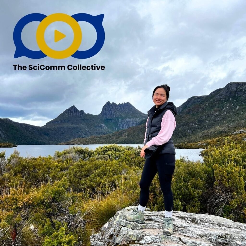 A woman standing on a rock with mountains and a lake behind her. The SciComm Collective logo of two speech bubbles and a play sign in a circle is on the top left.