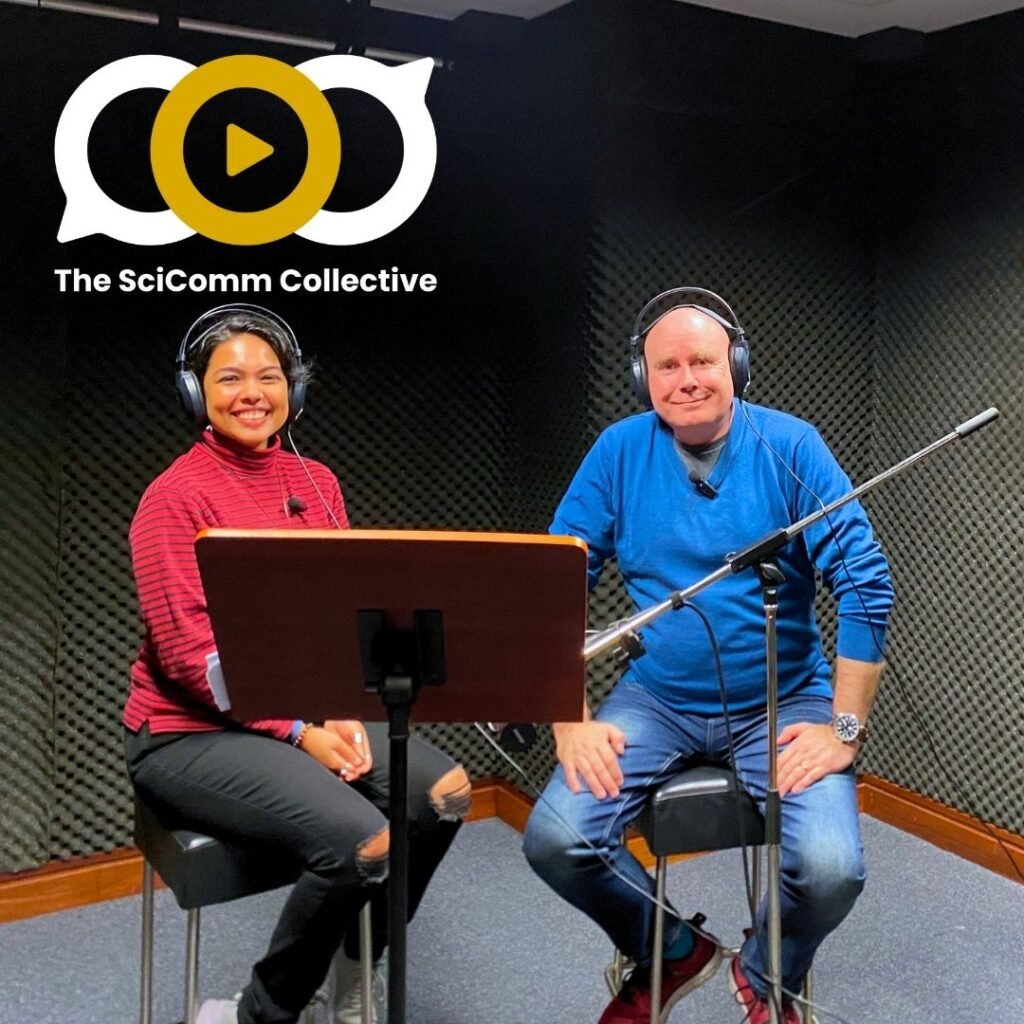 A smiling, bald white man in a blue jumper and a smiling younger Indian woman in a red jumper, both on stools wearing headphones, with a mic and a podium, in an audio studio