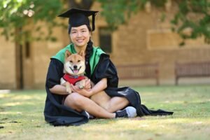 A smiling woman sitting cross-legged in a graduation gown on a lawn, holding her shiba inu