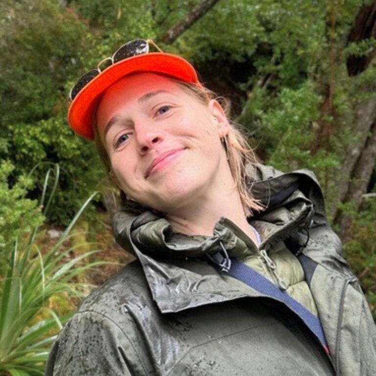 Young white woman with blond hair, a red cap, and green waterproof coat, outside, smiling at the camera with head tilted