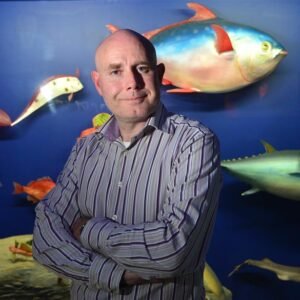 A white bald man with a striped shirt, arms crossed, smiling looking at the camera. Fish decorate the wall behind him