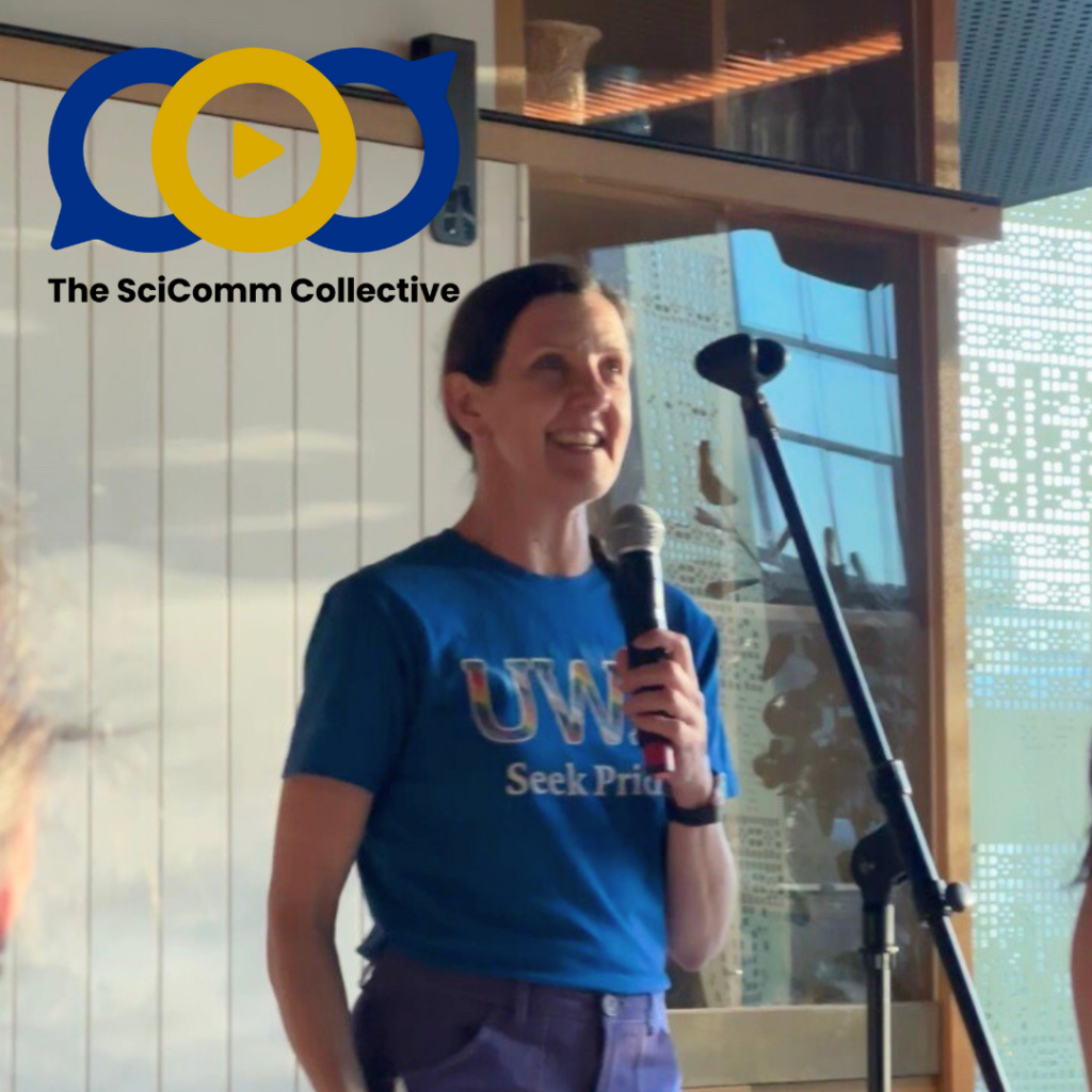 White woman with brown hair tied back, wearing a blue UWA Seek Pride tshirt and holding a microphone, smiling
