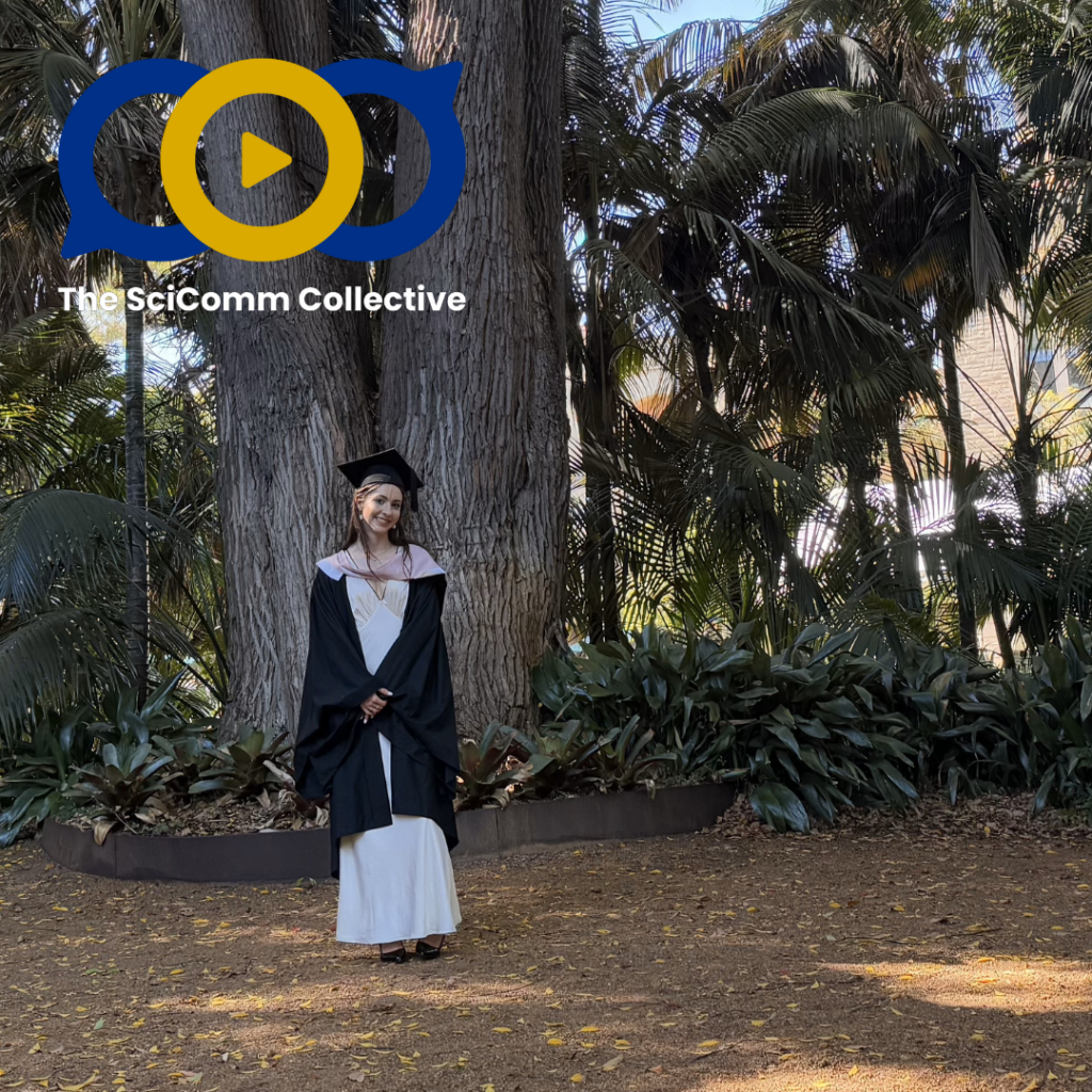 A smiling white woman with blonde hair and a white dress in a graduation gown and hat, standing before a tall tree in a shaded grove