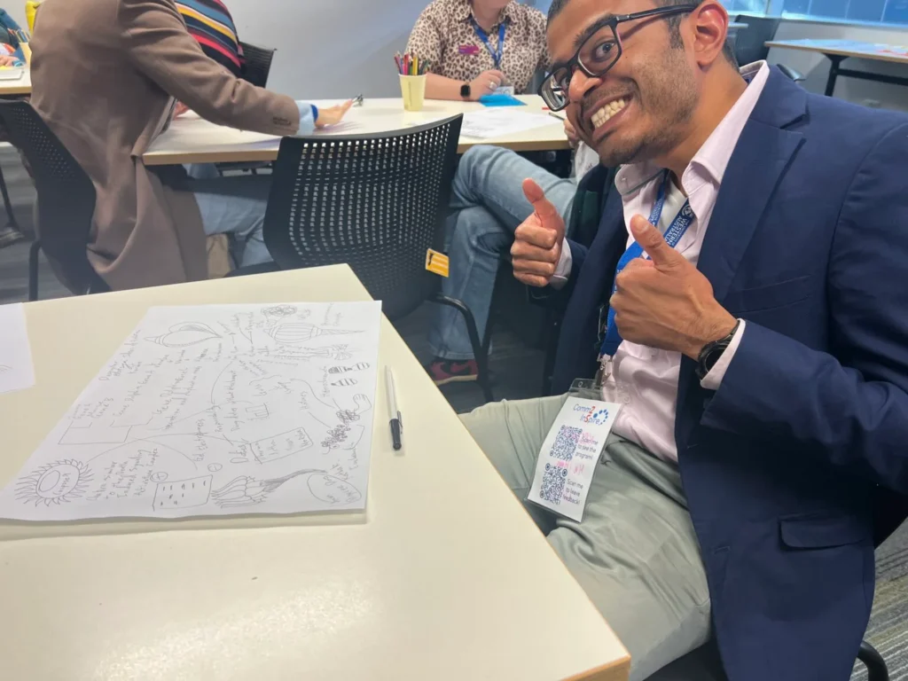 A man wearing glasses, a conference lanyard, jacket and smart attire is sitting and grinning with a head tilt and two thumbs up at the camera. On the table in front of him is a sheet of A3 paper, pen, and a drawn diagram which is unclear. More people sit at another table behind him.