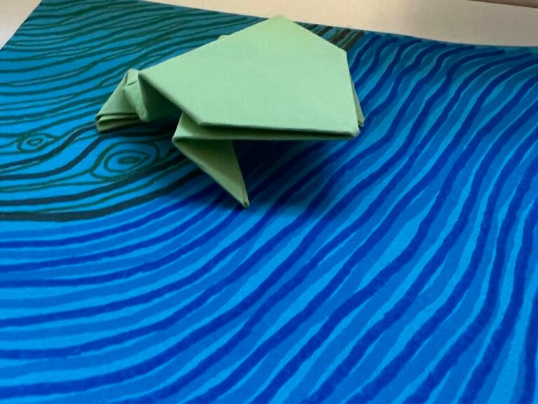 an origami frog made of green paper sitting on blue paper with waves drawn on it topographically