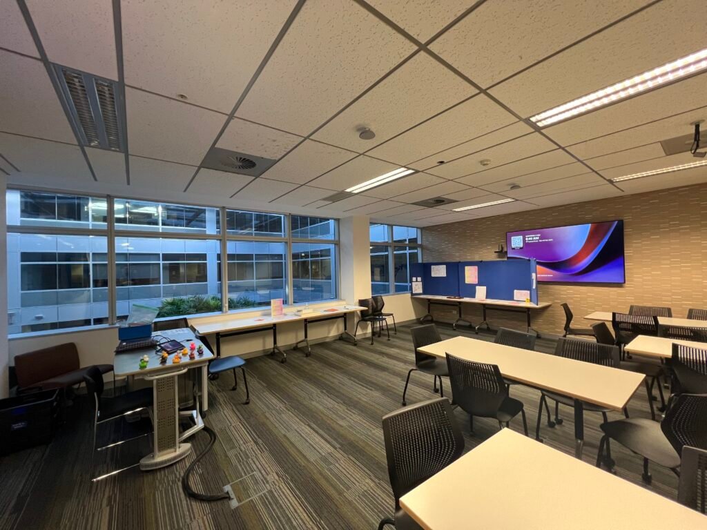 Photograph of a classroom with more buildings outside, chairs and tables arranged in groups, a blue board on a table at the back and a desk and chair at the front.
