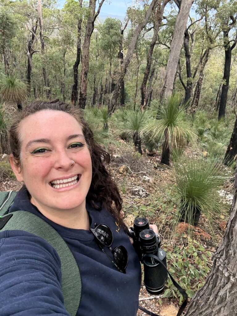 A selfie of a white woman smiling at the camera while holding a pair of black binoculars, wearing a jumper, backpack, and sunglasses on her collar, and with Australian bushland behind her.