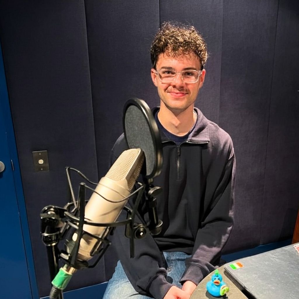 A 20-something white male with glasses, black zipper jumper and jeans, smiles at the camera while sitting in a soundproof recording studio, a mic on a stand nearby and a rubber duck and laptop on the table next to him
