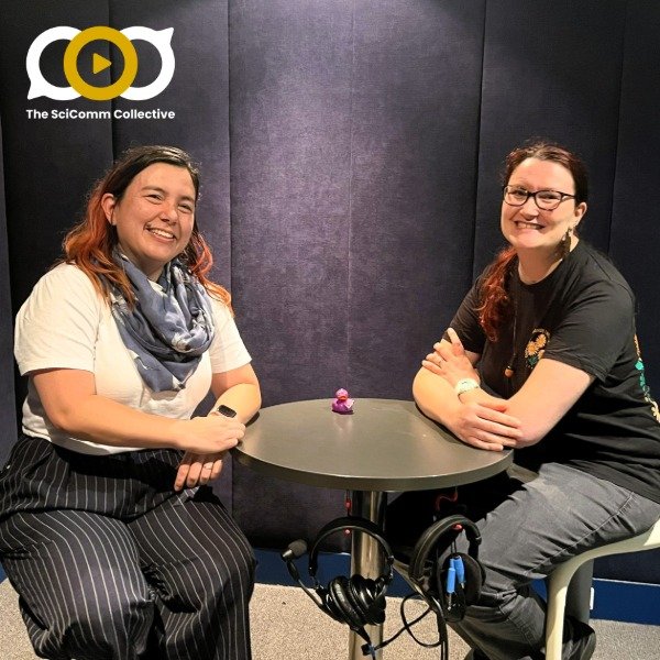 Two white women seated at a round table in a recording studio with dark purple walls and headsets hanging from the table; there is a small rubber duck on the table; the women are smiling at the camera. The SciComm Collective logo is on the image