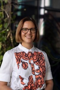 A white woman with a light brown bob and round dark glasses smiles at the camera; there is foliage in the background. She is wearing a collared shirt in white with an orange pattern design on the front.