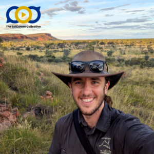 White man in a wide-brimmed hat and long-sleeved black shirt with a logo on his chest and sunglasses on the hat, taking a selfie in front of an open field landscape of natural scrub, low bushes and trees, and a distant red tabletop rise
