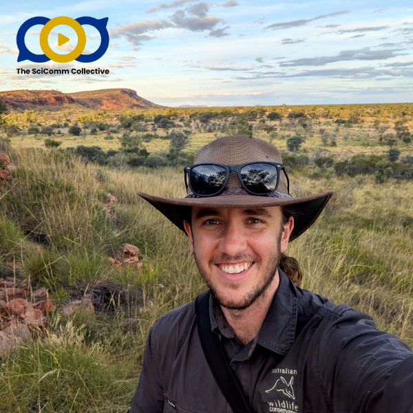 White man in a wide-brimmed hat and long-sleeved black shirt with a logo on his chest and sunglasses on the hat, taking a selfie in front of an open field landscape of natural scrub, low bushes and trees, and a distant red tabletop rise